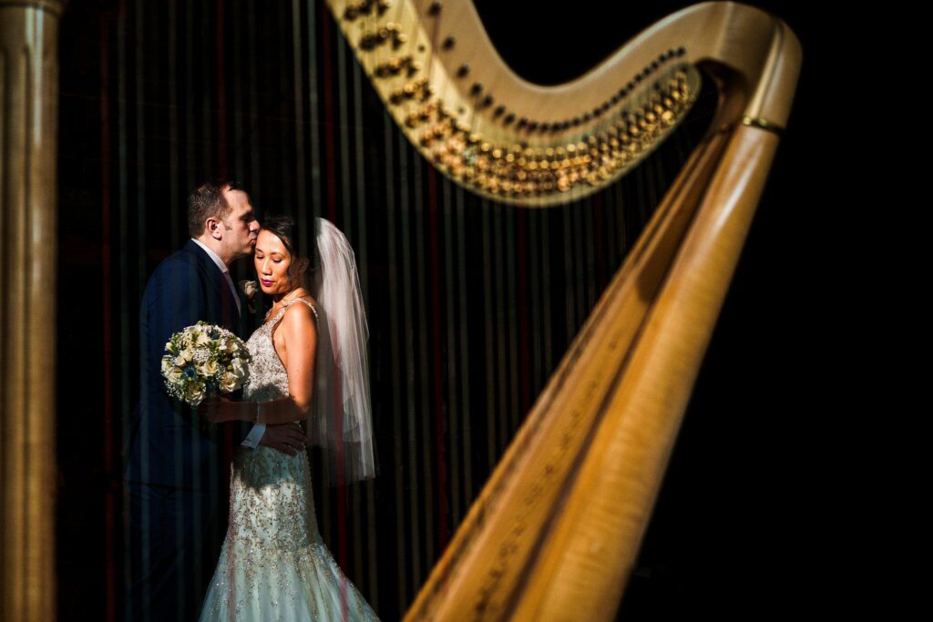 A bride and groom share an intimate moment as the groom whispers into the bride's ear. The bride holds a bouquet and wears a veil and a wedding dress. The image is framed by the strings of a harp in the foreground, adding a unique artistic touch. Image by Documentary Wedding Photographer Lewis Bishop Photography.