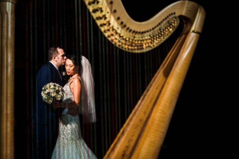 A bride and groom share an intimate moment, framed by the strings of a harp in the foreground. The groom is kissing the bride's forehead while she holds a bouquet of flowers and they are both dressed in wedding attire. The background is dark, highlighting the couple. Image by Documentary Wedding Photographer Lewis Bishop Photography.