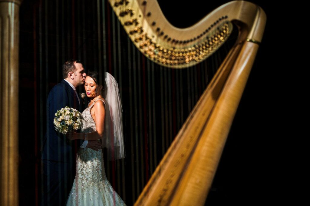 A bride in a white wedding gown holding a bouquet stands beside a groom in a dark suit, who is gently kissing her head. They are framed by the strings of a large harp in the foreground, creating an intimate and artistic composition. Image by Documentary Wedding Photographer Lewis Bishop Photography.