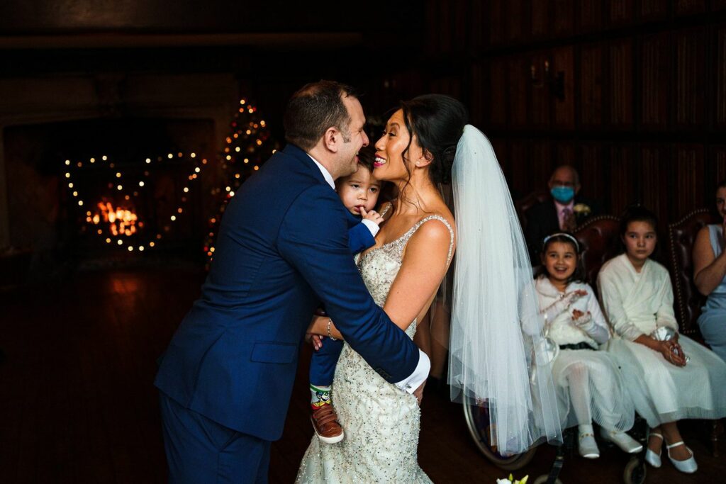 A bride and groom share a joyful moment indoors, with the groom holding a child. The bride is wearing a veil and a white dress, and the groom is in a blue suit. Behind them, a warm fireplace is lit with holiday decorations, and guests, including children, look on happily. Image by Documentary Wedding Photographer Lewis Bishop Photography.
