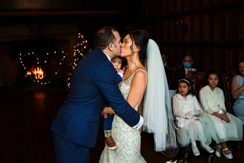 A bride and groom share a kiss during their wedding ceremony, holding a young child between them. The scene is set in a warmly lit room with a fireplace and Christmas tree in the background. Guests, including some children, are seated on the right. Image by Documentary Wedding Photographer Lewis Bishop Photography.