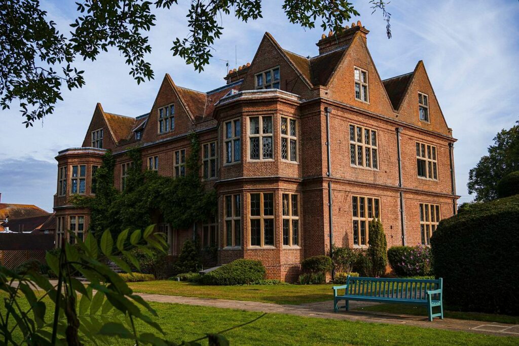 A large, historic brick building with multiple gabled roofs and numerous windows stands on a well-manicured lawn. A light blue wooden bench is situated in the foreground, surrounded by greenery and trees. The sky is clear, creating a picturesque scene. Image by Documentary Wedding Photographer Lewis Bishop Photography.