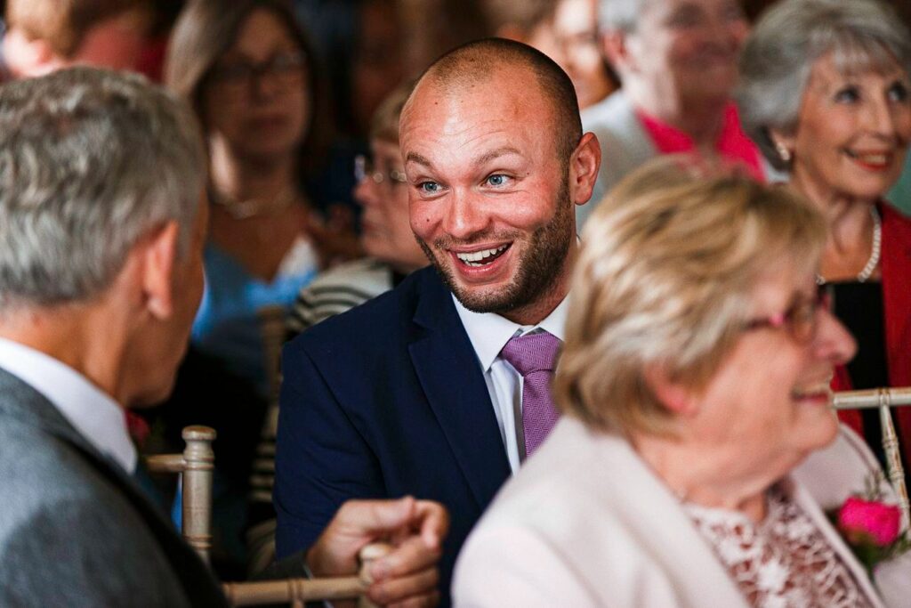 A bald man with a beard, wearing a dark blue suit and pink tie, is smiling and talking with an older gentleman at a social event. Other attendees, some sitting and some blurred in the background, are also seen conversing and enjoying themselves. Image by Documentary Wedding Photographer Lewis Bishop Photography.
