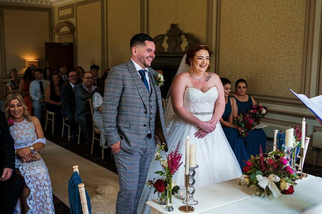 A bride in a white gown and a groom in a checked suit stand smiling near a table with candles and flowers, during a wedding ceremony. Seated guests in formal attire look on in the background, within a decorated room. Image by Documentary Wedding Photographer Lewis Bishop Photography.