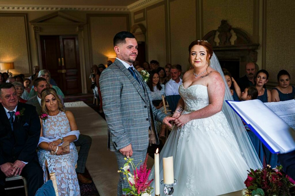 A bride and groom are standing and holding hands during their wedding ceremony in a decorated room filled with seated guests. The bride is wearing a white gown and veil, while the groom is in a gray plaid suit. A candle and flowers are visible in the foreground. Image by Documentary Wedding Photographer Lewis Bishop Photography.