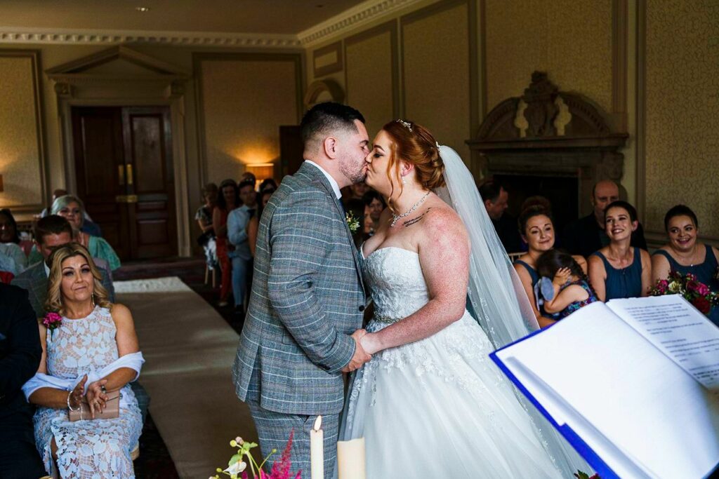 A newlywed couple kisses at the altar during their wedding ceremony. The groom is in a gray suit, and the bride is in a strapless white gown with a veil. Guests seated on either side watch the moment, and an official in the foreground holds the ceremony script. Image by Documentary Wedding Photographer Lewis Bishop Photography.