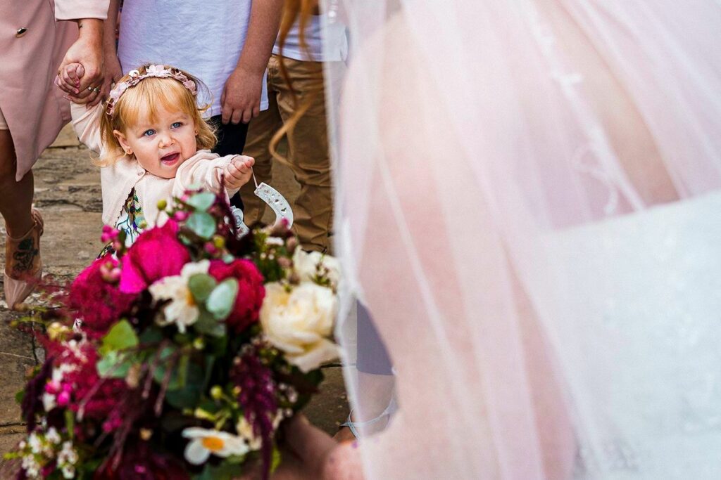 A young girl with light hair and a flower crown excitedly reaches out towards a bride in a white dress who is holding a vibrant bouquet of flowers. The background includes a couple of adults, partially visible, and the outdoor setting suggests a wedding scene. Image by Documentary Wedding Photographer Lewis Bishop Photography.