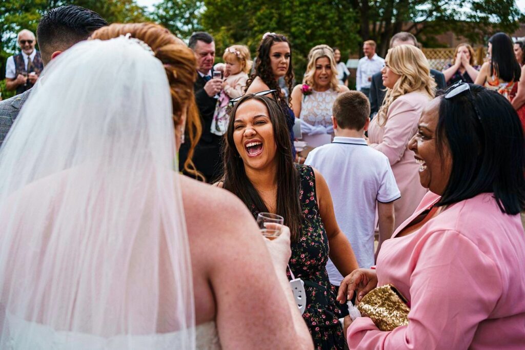 A bride in a white dress and veil stands with her back to the camera, engaging with guests at an outdoor wedding reception. One woman in a black dress laughs heartily, and another in a pink outfit smiles widely, holding a gold clutch. Other guests are in the background. Image by Documentary Wedding Photographer Lewis Bishop Photography.