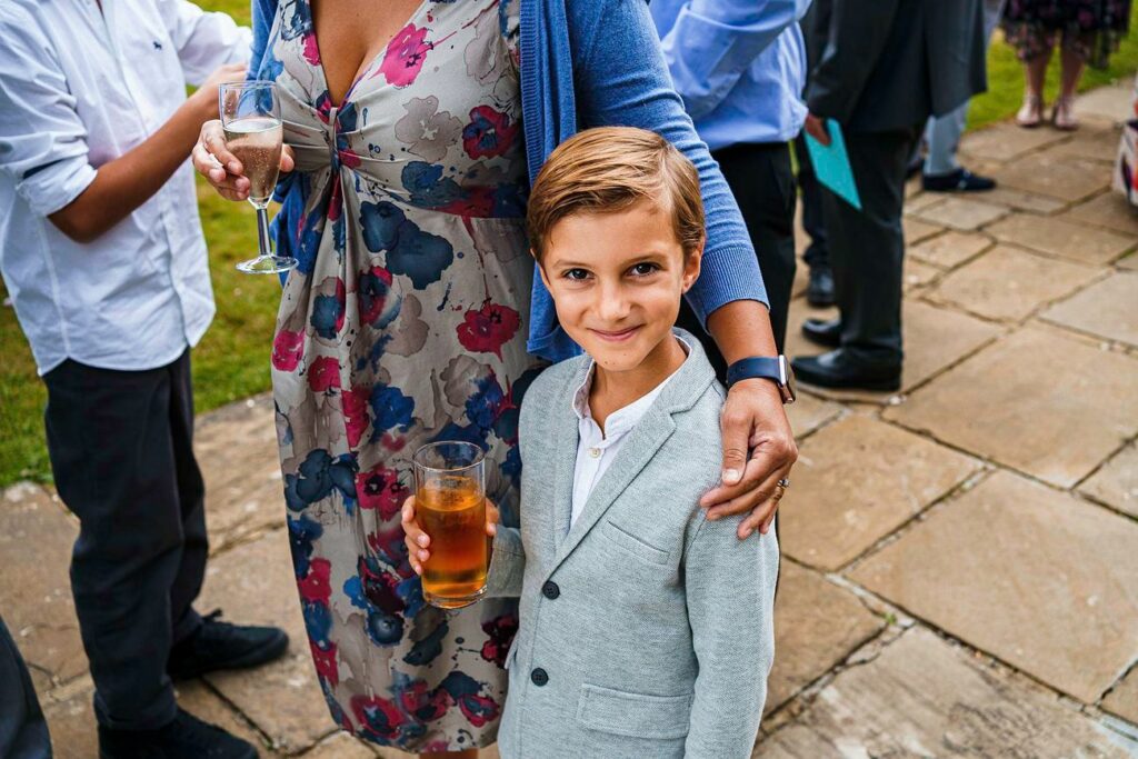 A young boy in a light gray suit stands smiling while holding a glass of juice. An adult woman, wearing a blue cardigan and floral dress, has her arm around his shoulders. They are outdoors on a stone patio along with other people who are partially visible. Image by Documentary Wedding Photographer Lewis Bishop Photography.