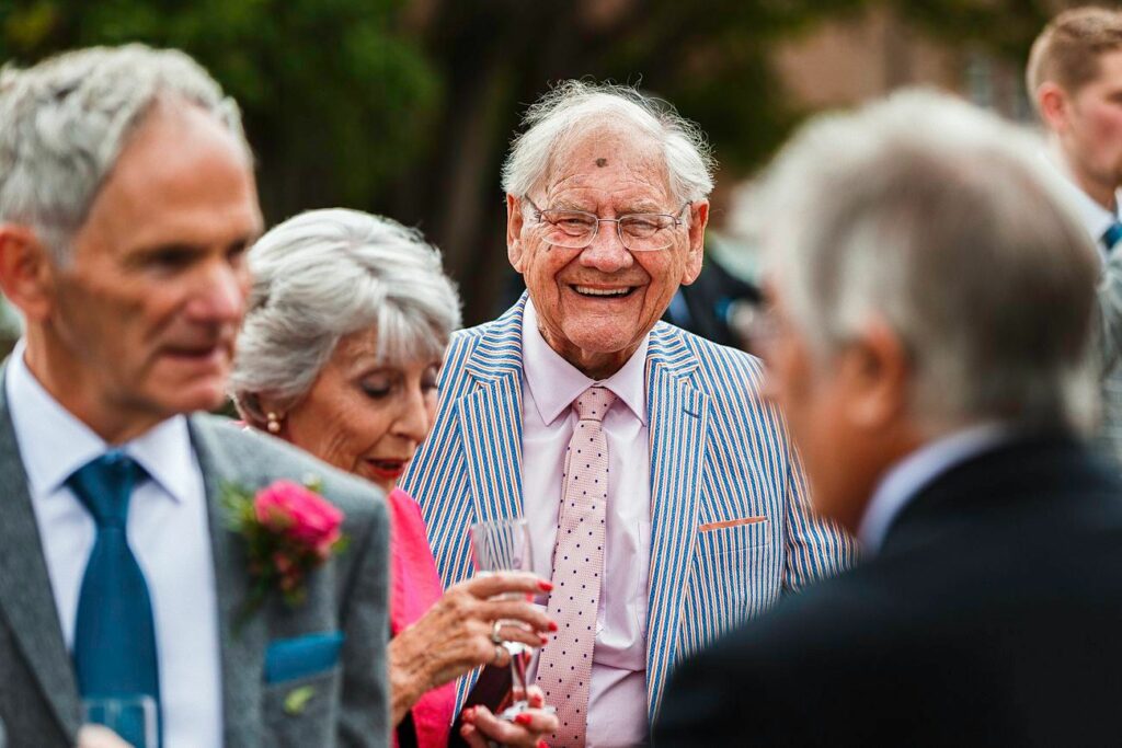 A smiling elderly man in a blue-striped suit and pink tie at an outdoor gathering. He stands among other formally dressed individuals, including a woman holding a drink and a man with a boutonniere. Lush green foliage is blurred in the background. Image by Documentary Wedding Photographer Lewis Bishop Photography.