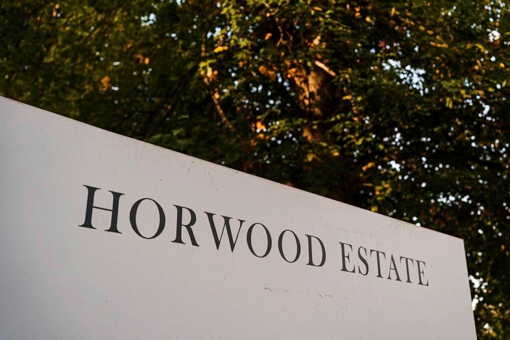A white sign with black text that reads "HORWOOD ESTATE" stands prominently in the foreground. In the background, there is lush green foliage and trees softly illuminated by sunlight. Image by Documentary Wedding Photographer Lewis Bishop Photography.