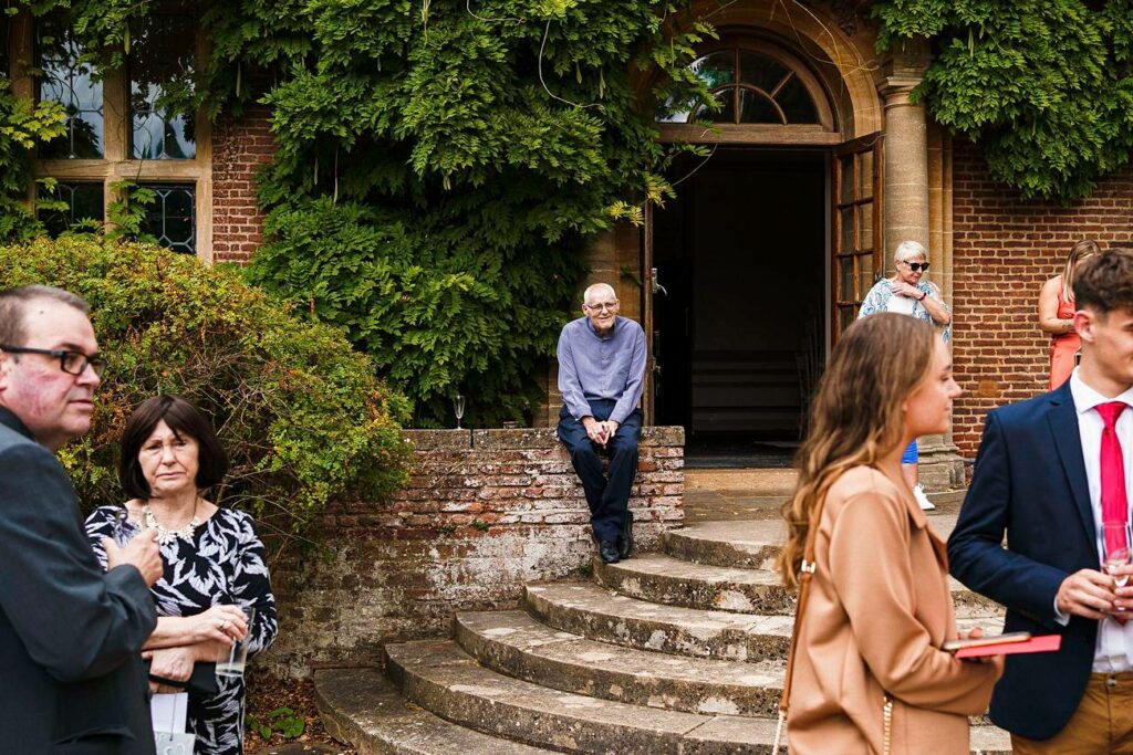 An elderly man wearing a sweater sits on the steps of a building with greenery. Others, dressed formally, are gathered nearby, one woman looking at the camera and others conversing. The scene suggests a social event in an outdoor setting. Image by Documentary Wedding Photographer Lewis Bishop Photography.