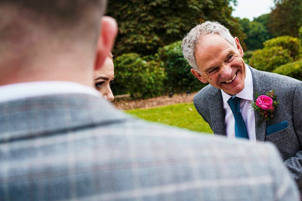An older man with gray hair and a big smile, wearing a gray suit with a bright pink boutonniere, stands outdoors. He is engaged with two other people, partially out of view. Greenery and trees are visible in the background. Image by Documentary Wedding Photographer Lewis Bishop Photography.