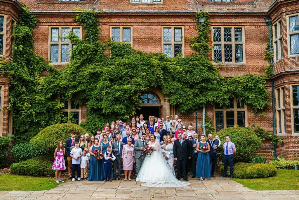 A large group of people pose for a wedding photo in front of an ivy-clad brick building with large windows. The bride and groom stand at the center, with bridesmaids in blue dresses and groomsmen in suits. Family and friends surround them, filling the steps and courtyard. Image by Documentary Wedding Photographer Lewis Bishop Photography.