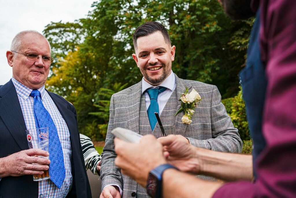 A man in a gray plaid suit with a blue tie and boutonniere is smiling and speaking with another person holding a phone. To his left, an older man in a navy suit with a patterned blue tie and glasses holds a drink. They are outdoors with greenery in the background. Image by Documentary Wedding Photographer Lewis Bishop Photography.