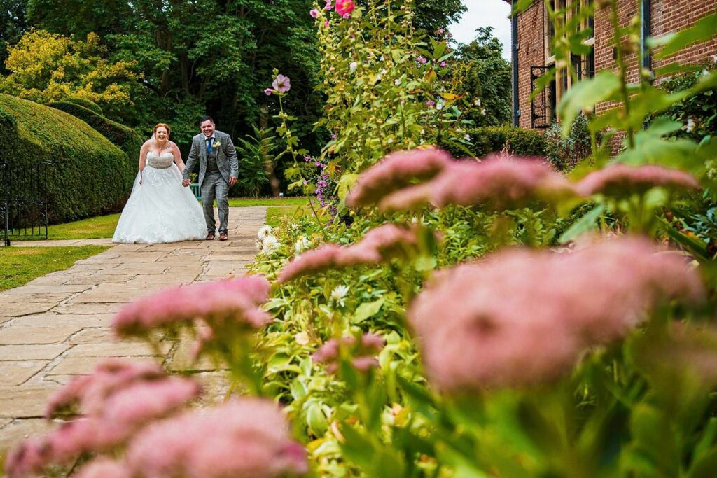 A bride and groom, holding hands, walk along a stone path through a garden filled with pink flowers and greenery. The bride wears a white wedding dress, and the groom is in a grey suit. The path borders a brick building, and trees are visible in the background. Image by Documentary Wedding Photographer Lewis Bishop Photography.