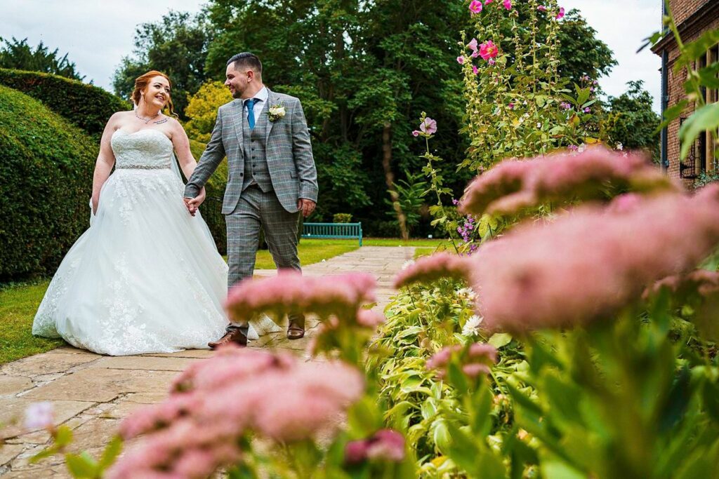 A bride in a white gown and a groom in a grey plaid suit walk hand-in-hand along a garden pathway lined with pink flowers. They are smiling at each other, with a background of lush green trees and a wooden bench. Image by Documentary Wedding Photographer Lewis Bishop Photography.