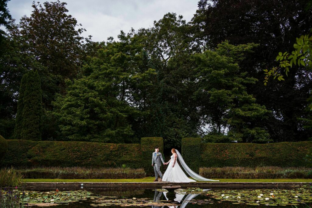 A bride and groom hold hands and walk along a path beside a lily pond in a lush garden. The bride wears a flowing white gown with a long veil, and the groom wears a light gray suit. Tall, green hedges and trees form the backdrop under an overcast sky. Image by Documentary Wedding Photographer Lewis Bishop Photography.