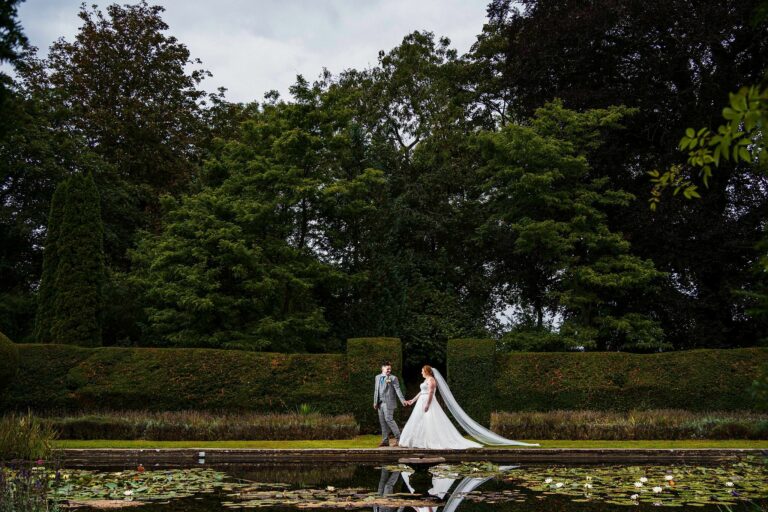 A bride and groom hold hands and walk along a path beside a lily pond in a lush garden. The bride wears a flowing white gown with a long veil, and the groom wears a light gray suit. Tall, green hedges and trees form the backdrop under an overcast sky. Image by Documentary Wedding Photographer Lewis Bishop Photography.