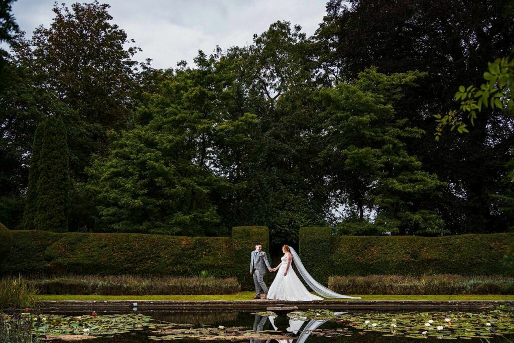 A bride and groom hold hands and walk near a serene pond with lily pads, surrounded by lush greenery and tall trees in an outdoor garden setting. The bride wears a white gown with a long veil, and the groom is dressed in a light gray suit. Image by Documentary Wedding Photographer Lewis Bishop Photography.