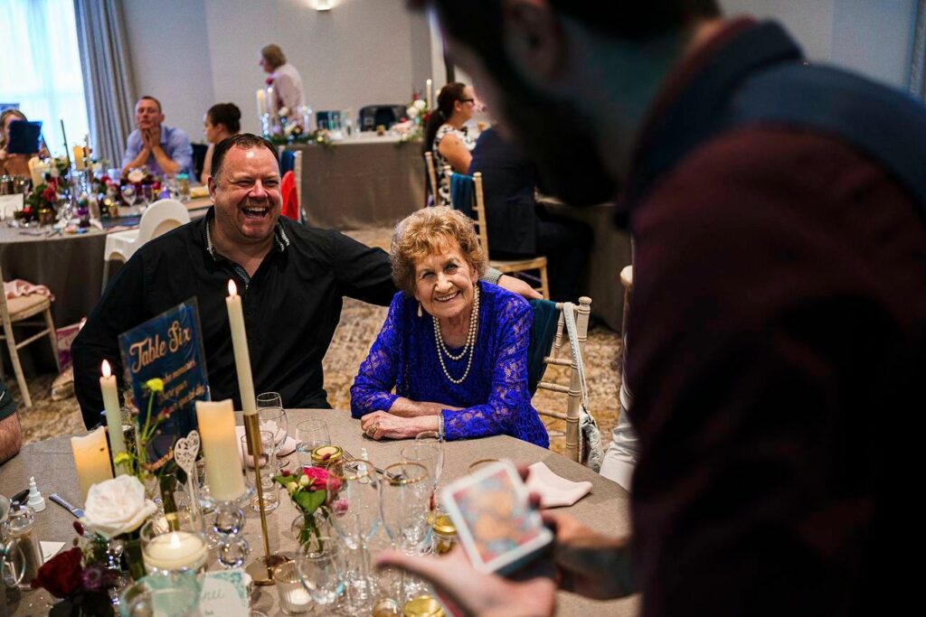 A joyous elderly woman and a man laugh together at a table adorned with flowers, candles, and glasses at a lively event. A person standing nearby performs a card trick, captivating their attention. Other guests are seated in the background, engaged in conversation. Image by Documentary Wedding Photographer Lewis Bishop Photography.