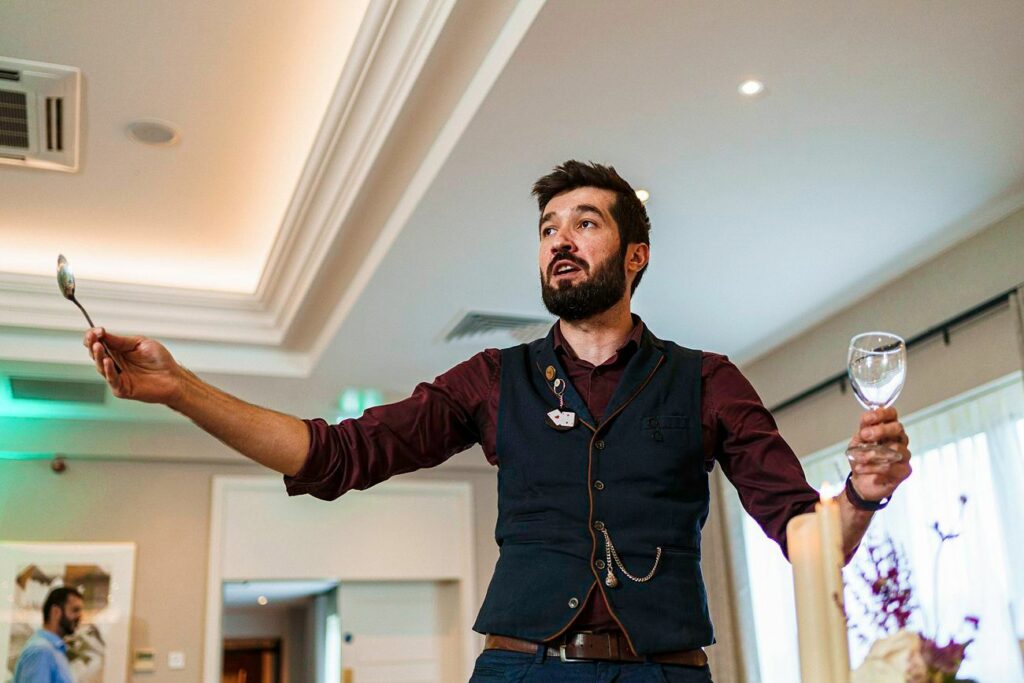 A man with a beard stands gesturing expressively with a spoon in one hand and an empty wine glass in the other. He is wearing a dark vest over a maroon shirt and appears to be speaking or making a presentation in a well-lit room. Image by Documentary Wedding Photographer Lewis Bishop Photography.