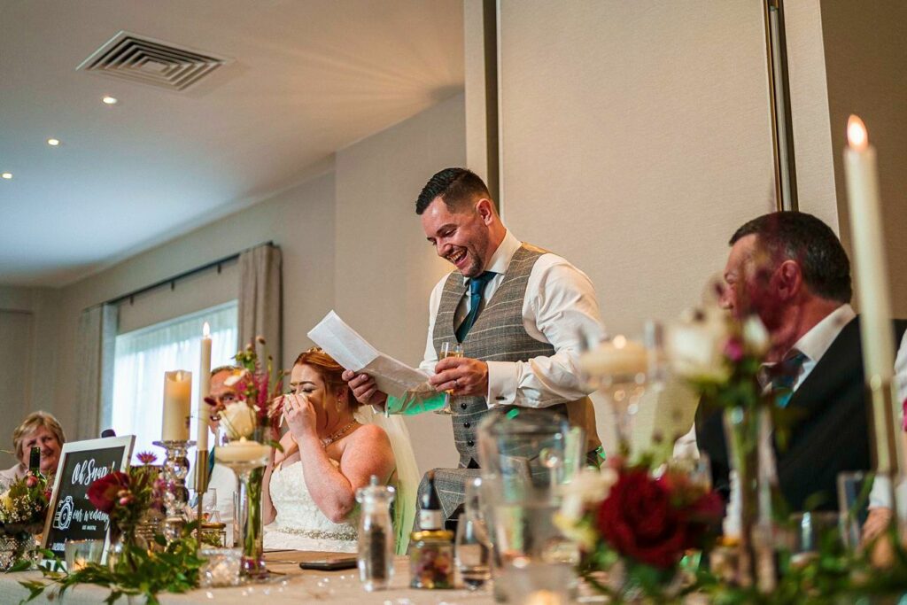 A man stands at a decorated table, reading from a piece of paper and smiling. A woman beside him, likely the bride, sits with her hand covering her mouth, appearing emotional. Other people are seated nearby, and the room is adorned with flowers and candles. Image by Documentary Wedding Photographer Lewis Bishop Photography.