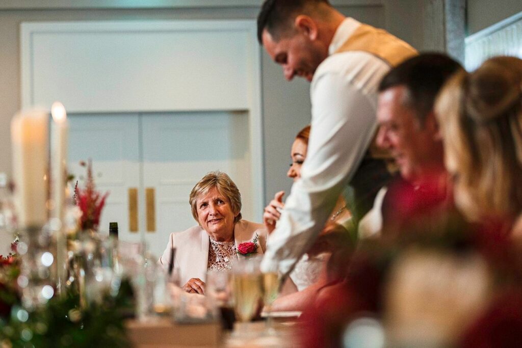 A group of people are seated at a table during a wedding reception. A man in a vest and tie stands up while speaking, looking down at a woman in a white dress beside him. One woman, wearing a pink outfit, smiles at the couple. The table is decorated with floral arrangements and candles. Image by Documentary Wedding Photographer Lewis Bishop Photography.