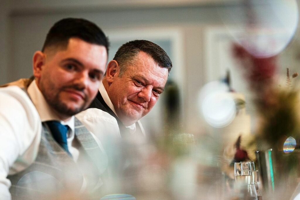 Two men sitting next to each other in a dimly lit room. The man on the left has short hair and a trimmed beard, and is wearing a white shirt with a gray vest. The older man on the right is smiling, wearing a dark vest over a white shirt. The background is blurred. Image by Documentary Wedding Photographer Lewis Bishop Photography.