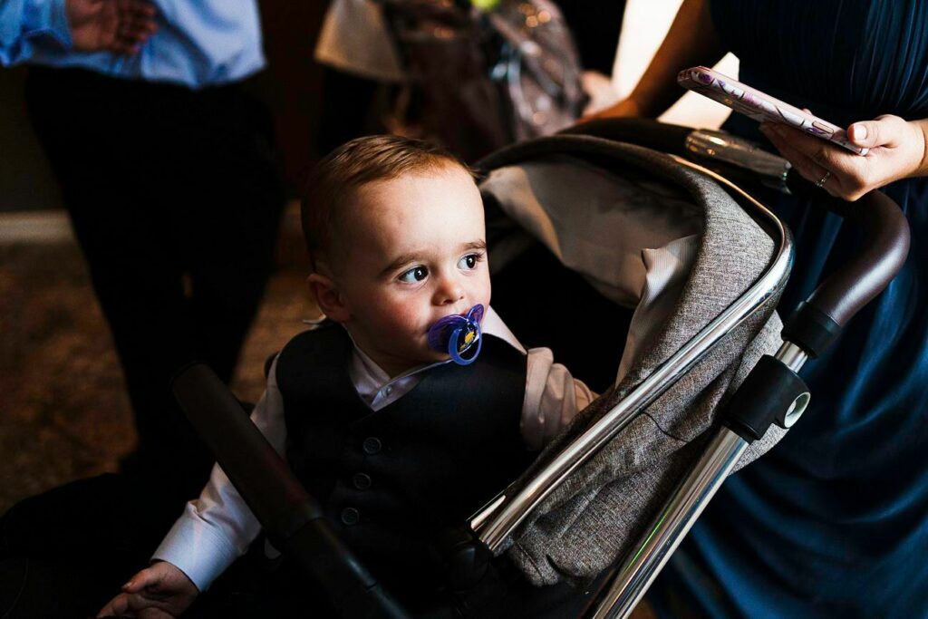 A toddler with short light brown hair sits in a stroller, wearing a white shirt and dark vest, and has a blue pacifier in his mouth. An adult beside him holds a smartphone, with only their hand visible. The background is dimly lit and slightly blurred. Image by Documentary Wedding Photographer Lewis Bishop Photography.