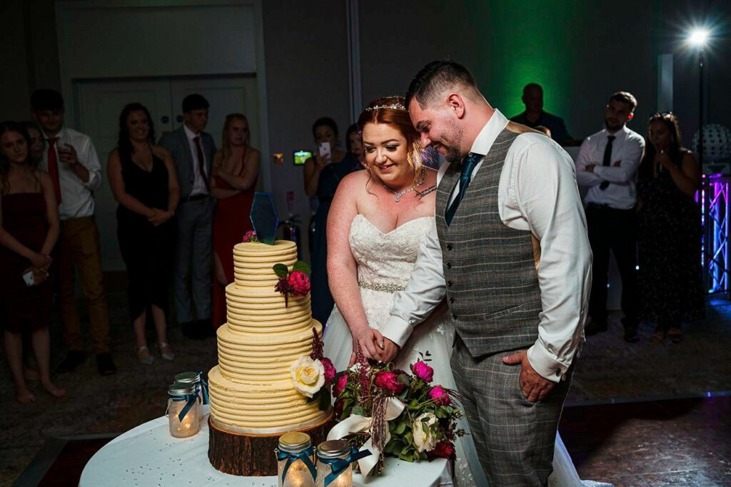 A bride and groom smile as they cut a large tiered wedding cake decorated with flowers on top. The bride wears a white gown, and the groom is in a gray plaid suit. They are surrounded by guests who are watching and smiling in the background. Image by Documentary Wedding Photographer Lewis Bishop Photography.