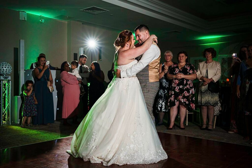 A couple in wedding attire shares a first dance on the dance floor, surrounded by smiling guests. The bride is wearing a white gown and the groom is in a vest and pants. Green lighting and a spotlight enhance the festive atmosphere. Image by Documentary Wedding Photographer Lewis Bishop Photography.