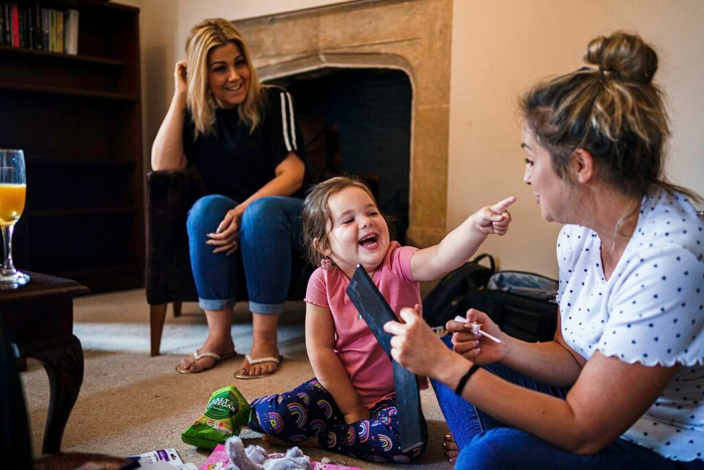 A young child, smiling and pointing excitedly, sits between two women in a cozy living room. One woman watches with a smile from a chair, while the other engages with the child, holding a toy. A glass of orange juice is on the table next to them. Image by Documentary Wedding Photographer Lewis Bishop Photography.
