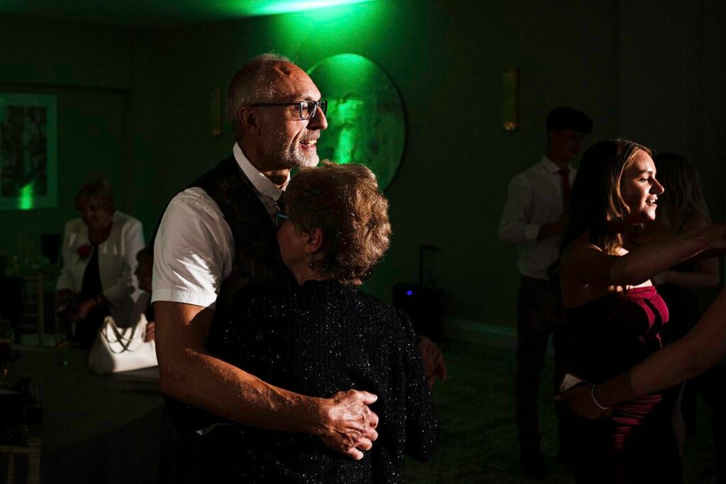 An elderly couple is seen dancing closely in a dimly lit room with a green spotlight highlighting them. The man is wearing glasses and a vest, and the woman is dressed in a glittery black outfit. Other people are dancing in the background, enjoying the occasion. Image by Documentary Wedding Photographer Lewis Bishop Photography.