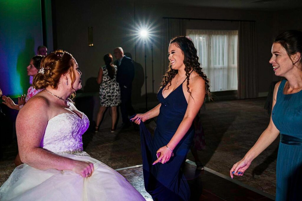 Three women are joyfully dancing at a wedding reception. The bride, in a white gown, and two bridesmaids, wearing dark blue and teal dresses, are enjoying the moment. In the background, there are other guests and a bright light illuminating the scene. Image by Documentary Wedding Photographer Lewis Bishop Photography.