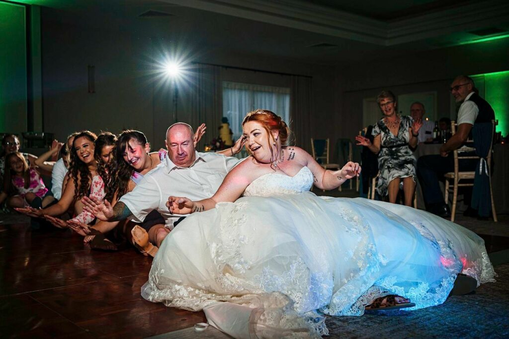 A joyful bride in a white wedding dress leads a group of guests, including an older man in a white shirt, in a dance move on the floor at a wedding reception. They appear to be laughing and having fun, while other guests watch and smile in the background. Image by Documentary Wedding Photographer Lewis Bishop Photography.