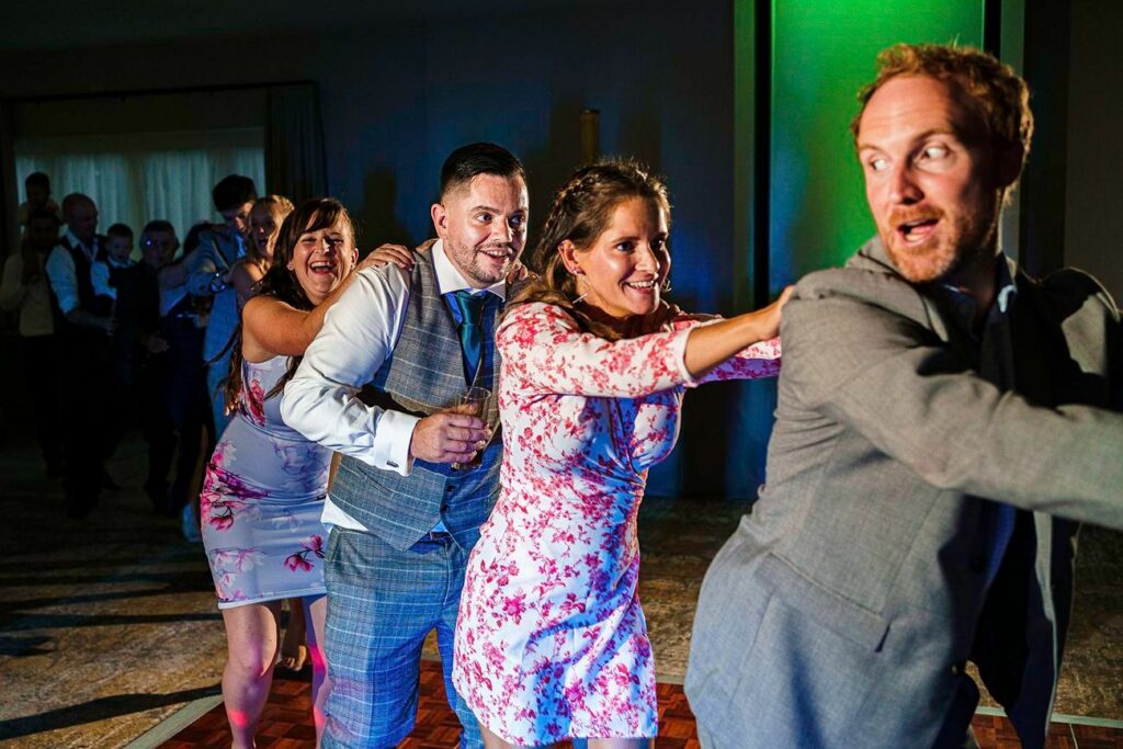 A group of people, dressed in formal attire, dance in a conga line at a party or celebration. The people are smiling and laughing, with their hands on each other's shoulders, illuminated by colorful, festive lighting. Image by Documentary Wedding Photographer Lewis Bishop Photography.