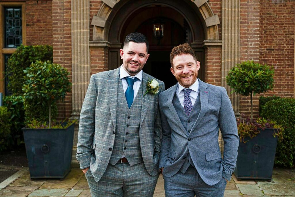 Two men stand side by side, smiling, dressed in suits. The man on the left is wearing a gray checkered three-piece suit with a white shirt and teal tie, while the man on the right is in a plain gray suit with a white shirt and patterned tie. They are standing in front of a brick building entrance framed by columns and greenery. Image by Documentary Wedding Photographer Lewis Bishop Photography.