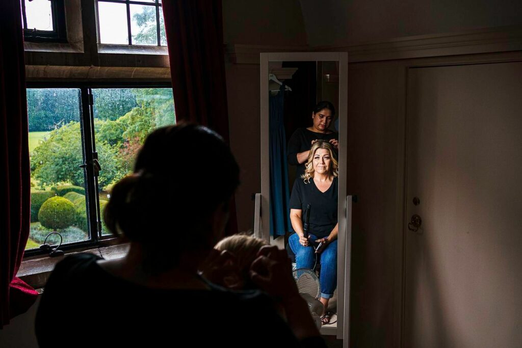 A woman in a black shirt sits in front of a tall mirror as another woman styles her hair. The scene is illuminated by natural light from a window with a view of lush green trees and bushes. Another person is seen in the foreground, adjusting a child's hair. Image by Documentary Wedding Photographer Lewis Bishop Photography.