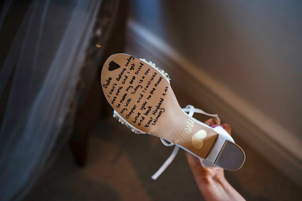 A close-up of the sole of a white shoe held in a hand. The sole has a handwritten message in black ink addressed to "Babe" with a heart symbol above the message. The background includes part of a veil and some furniture. Image by Documentary Wedding Photographer Lewis Bishop Photography.