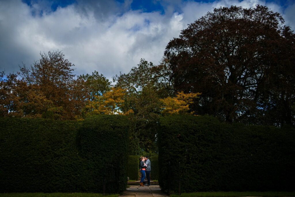 A couple stands closely together at the entrance of a large hedge maze, surrounded by tall, lush trees with autumn-colored leaves under a partly cloudy sky. The scene conveys a serene, romantic atmosphere. Image by Documentary Wedding Photographer Lewis Bishop Photography.