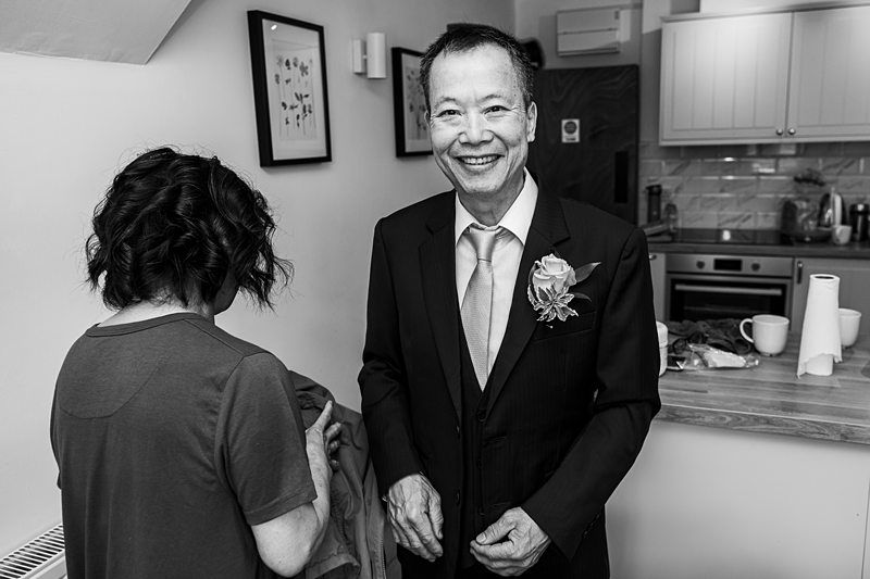 A man in a suit, wearing a boutonnière, smiles while a woman stands next to him, looking down at a garment. They are in a kitchen area with minimal decor. Image by Documentary Wedding Photographer Lewis Bishop Photography.