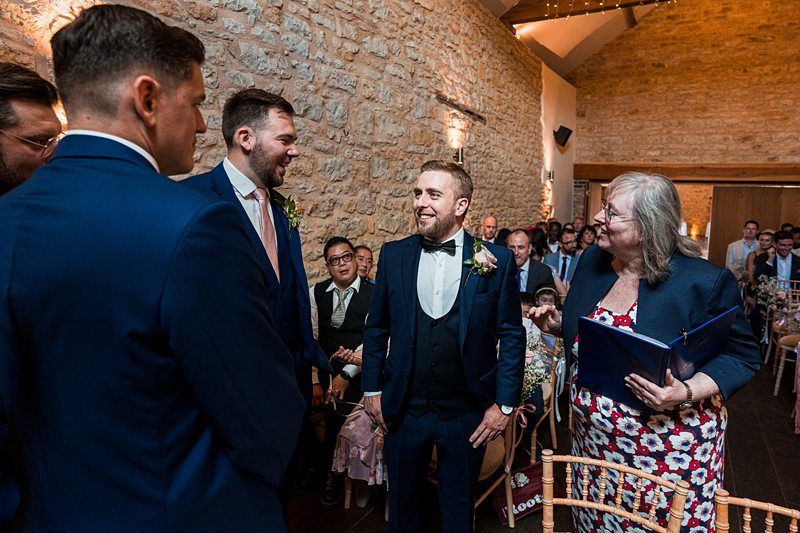 A group of people, including men in suits and a woman holding a blue folder, are smiling and interacting at a social gathering in a brick-walled, decorated venue. Image by Documentary Wedding Photographer Lewis Bishop Photography.