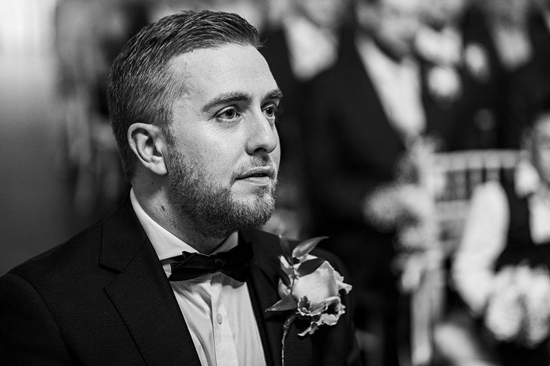A man in formal attire with a bow tie and flower boutonniere sits among people in similar attire. Image by Documentary Wedding Photographer Lewis Bishop Photography.