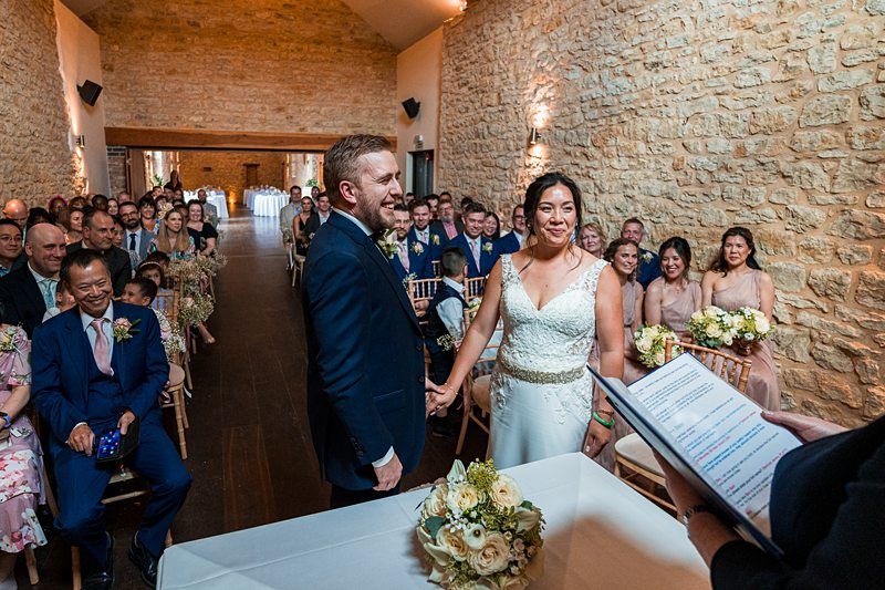 A couple stands at the altar during their wedding ceremony in a rustic stone-walled venue, surrounded by seated guests. The officiant holds a booklet with the ceremony details. Image by Documentary Wedding Photographer Lewis Bishop Photography.
