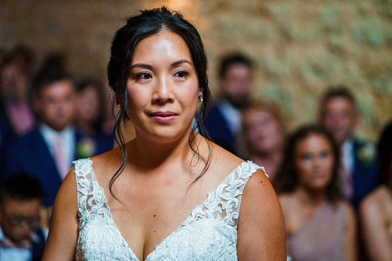 A woman in a white lace dress stands indoors, with a thoughtful expression. Blurred people are visible in the background. Image by Documentary Wedding Photographer Lewis Bishop Photography.