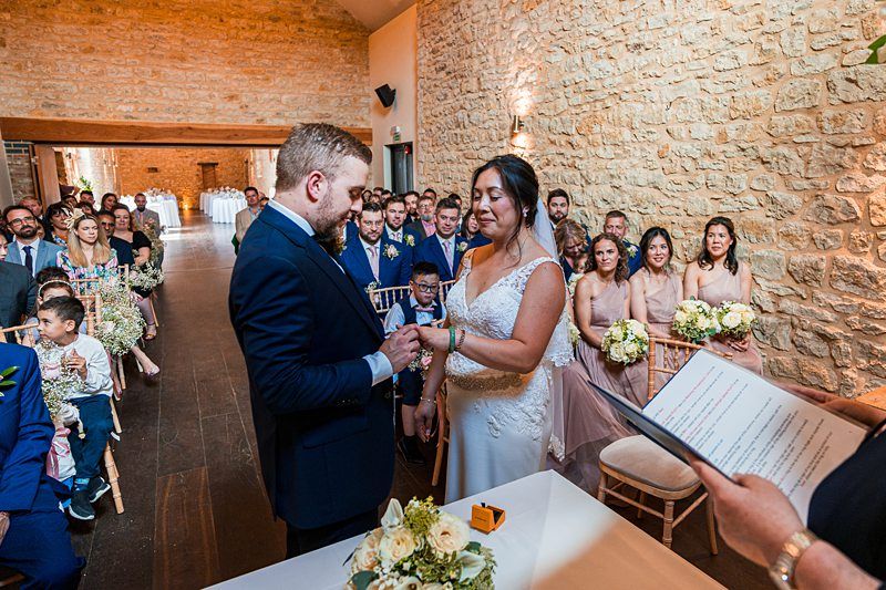 A couple stands facing each other, holding hands, during a wedding ceremony in a rustic stone-walled venue. Guests are seated and watching. An officiant holding a book stands nearby. Image by Documentary Wedding Photographer Lewis Bishop Photography.