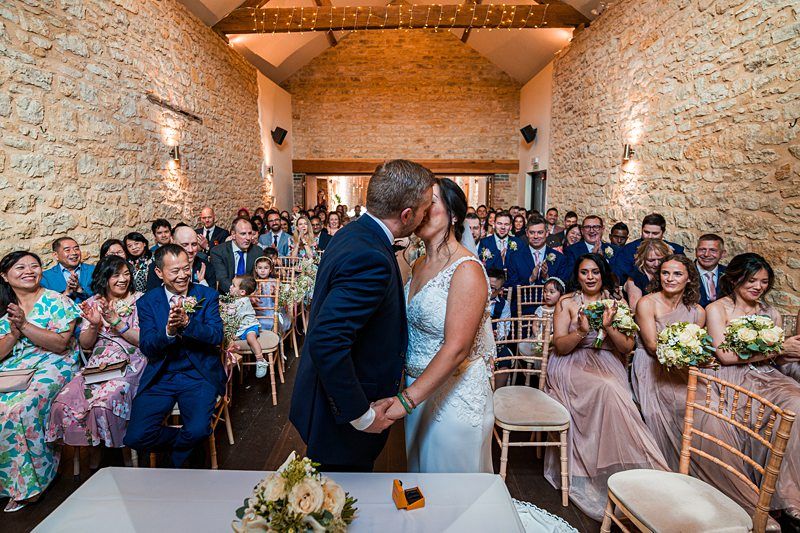 A bride and groom kiss at the altar in a rustic stone hall, surrounded by guests who are smiling and clapping. The wedding party is seated on both sides, holding floral bouquets. Image by Documentary Wedding Photographer Lewis Bishop Photography.
