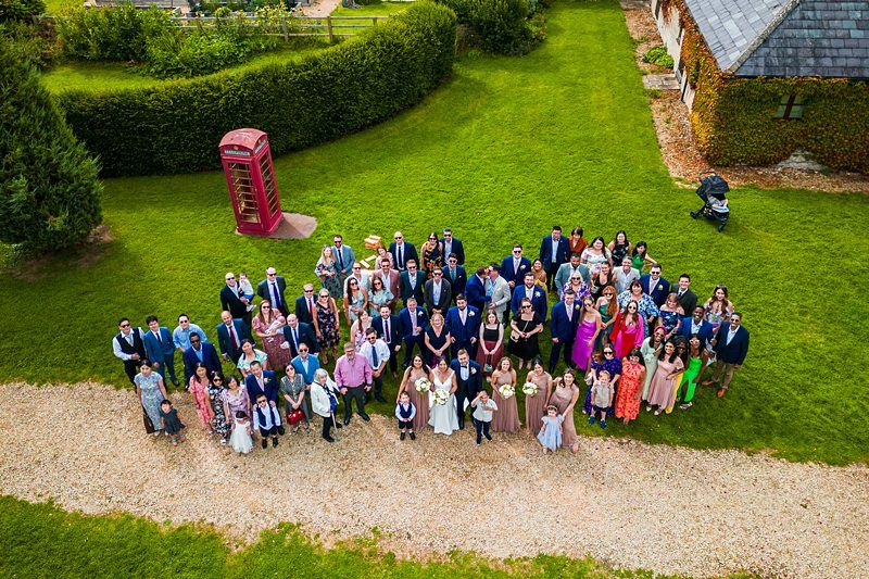A large group of people dressed formally, posing outdoors in a garden setting, with a red phone booth on the left and a building covered with ivy on the right. Image by Documentary Wedding Photographer Lewis Bishop Photography.
