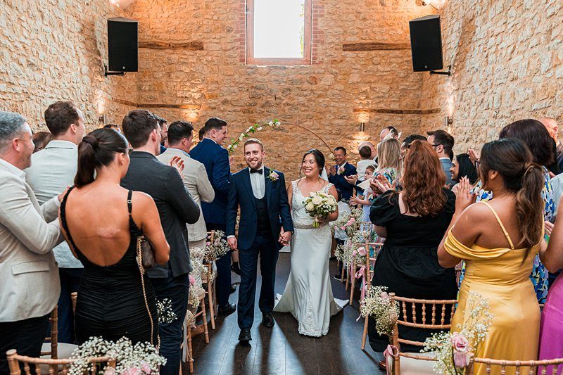 Bride and groom walk down the aisle in a stone-walled venue, greeted by standing guests. The bride holds a bouquet and both are smiling, surrounded by seated and standing attendees on either side. Image by Documentary Wedding Photographer Lewis Bishop Photography.